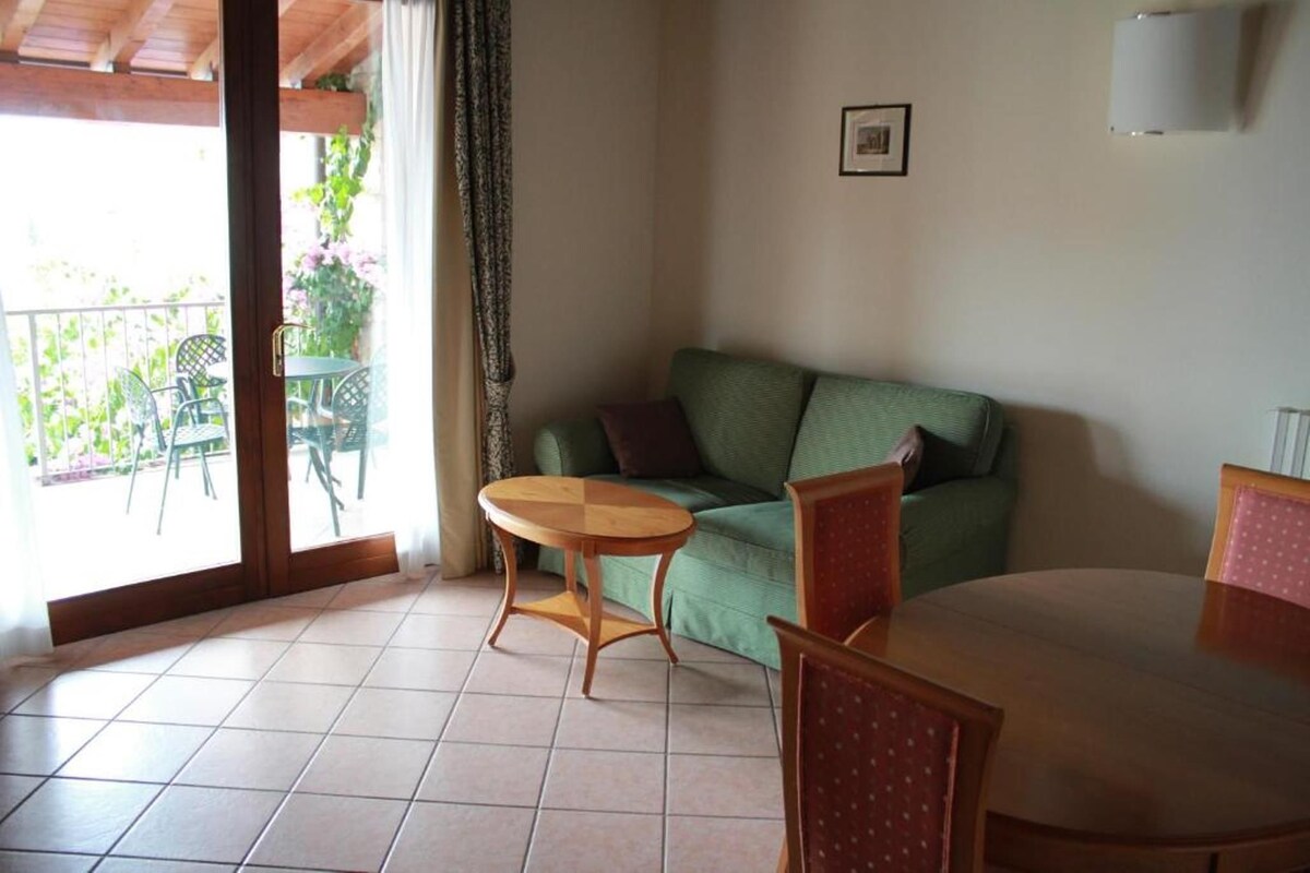 Una habitación con un sofá verde, una mesa de madera y una silla. Se ve una ventana con vistas a muebles de exterior y plantas.