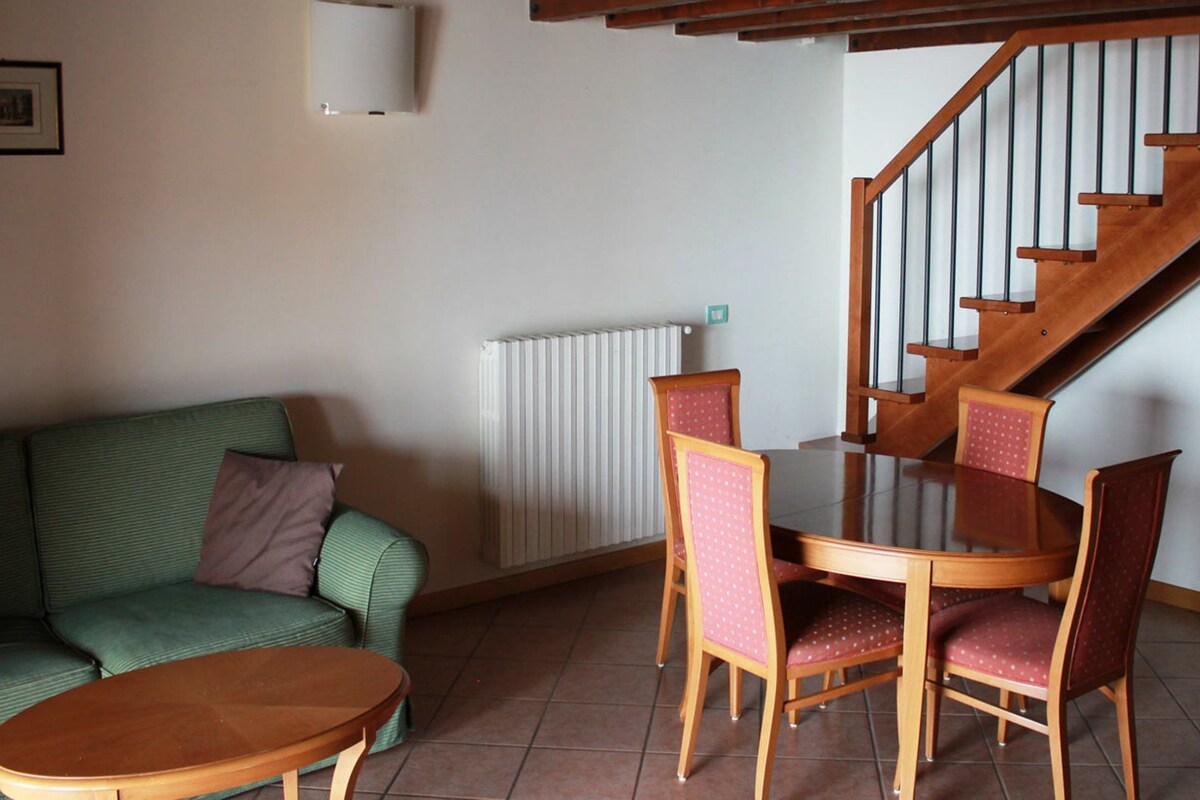Una sala de estar con un sofá verde, una mesa de centro de madera y una mesa de comedor con sillas. Se ve una escalera con barandillas de madera al fondo.