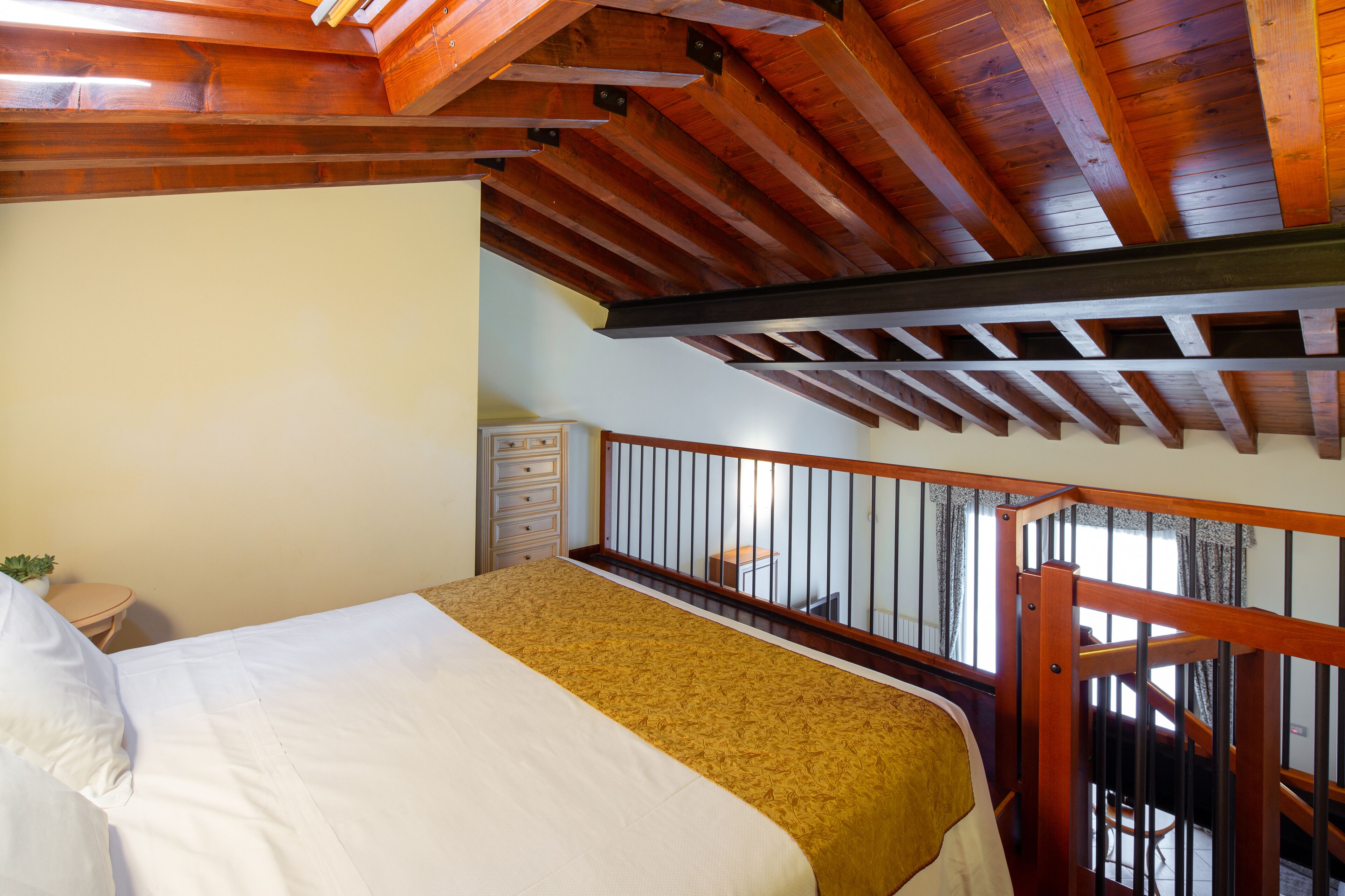 Une chambre avec un lit, des poutres en bois au plafond, un balcon avec une balustrade et une petite table avec une plante.