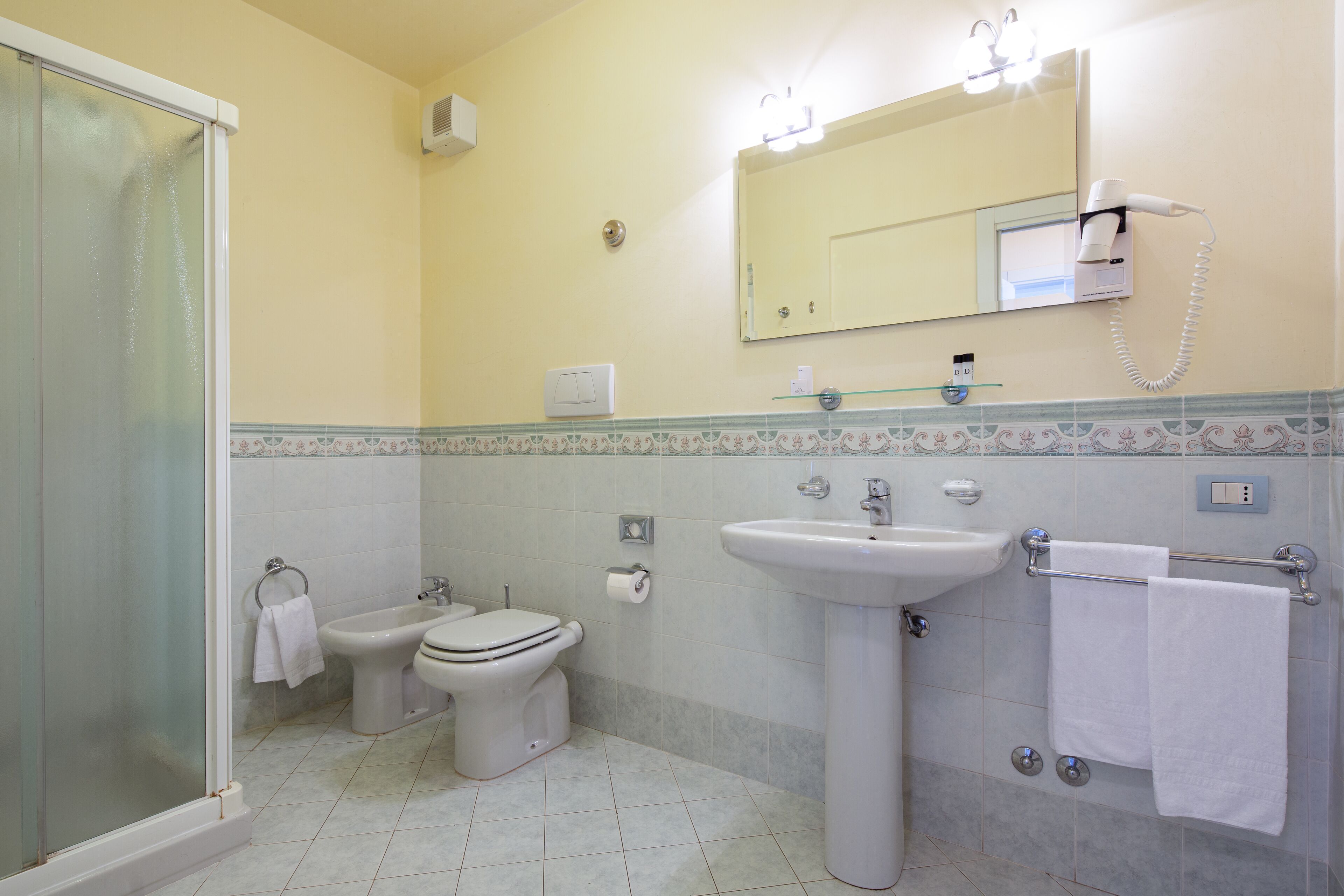 Une salle de bain équipée d’une douche, de toilettes, d’un bidet, d’un lavabo et d’un sèche-cheveux fixé au mur.