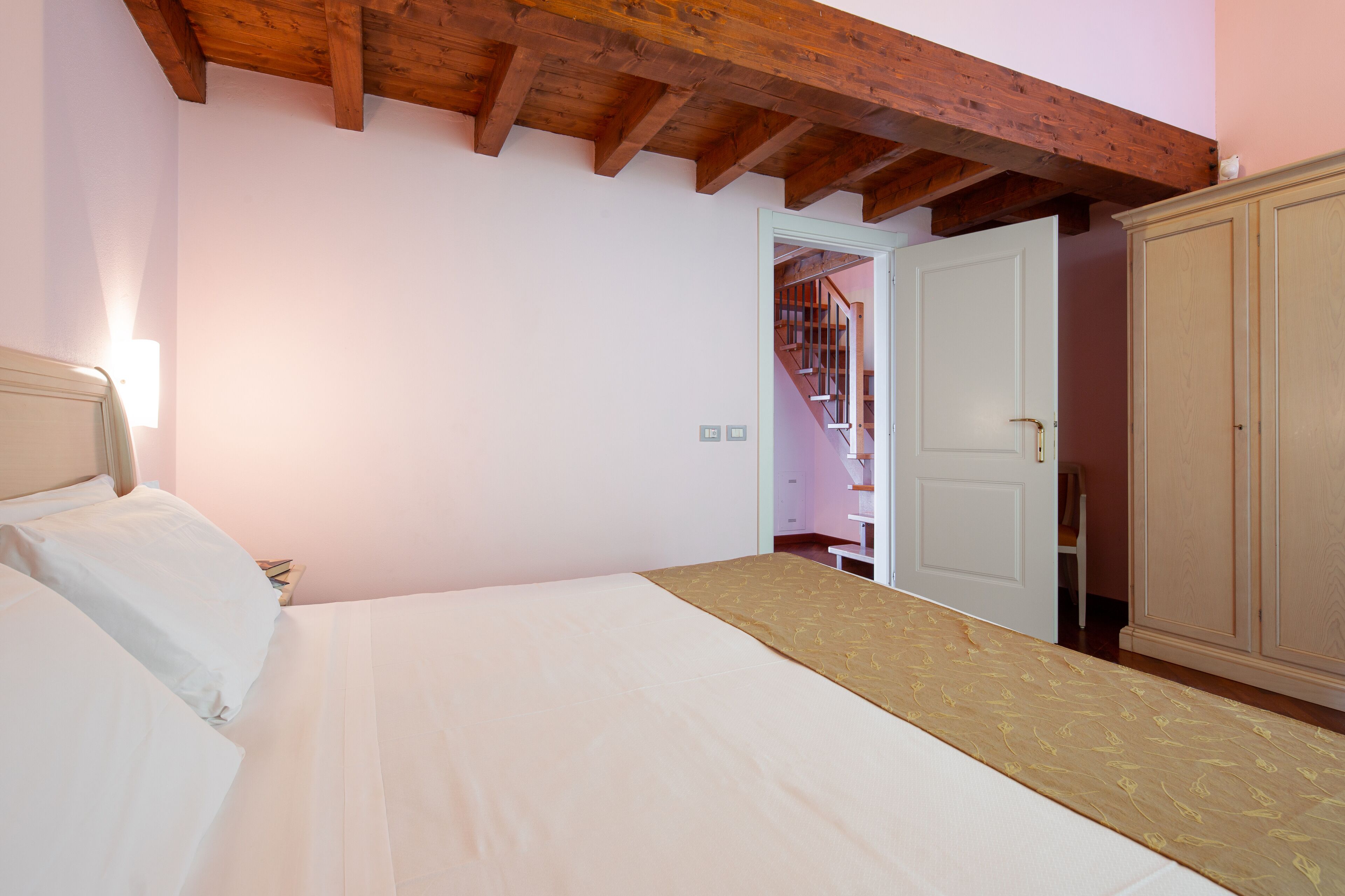 A bedroom with a bed, wooden ceiling beams, a wardrobe, and a staircase leading to another room.