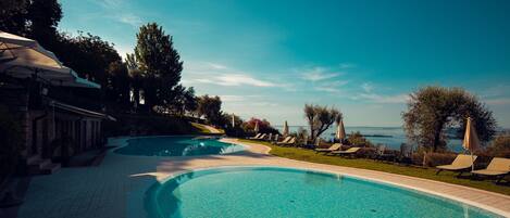 A swimming pool with a curved edge, surrounded by lounge chairs and umbrellas, with a view of the sea in the background.