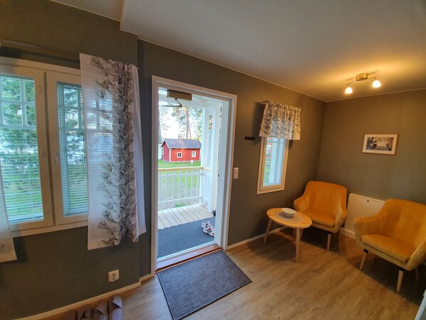 A room with a wooden floor, a small table with a chair, a door leading outside, and a window with curtains.