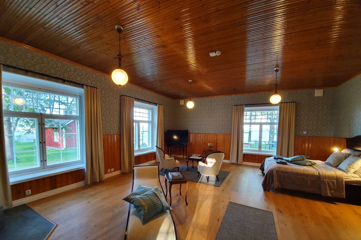 Ruime kamer met houten vloeren en een hoge plafond, met een bed, een bank, een eettafel met stoelen en grote ramen met uitzicht op het omliggende landschap.