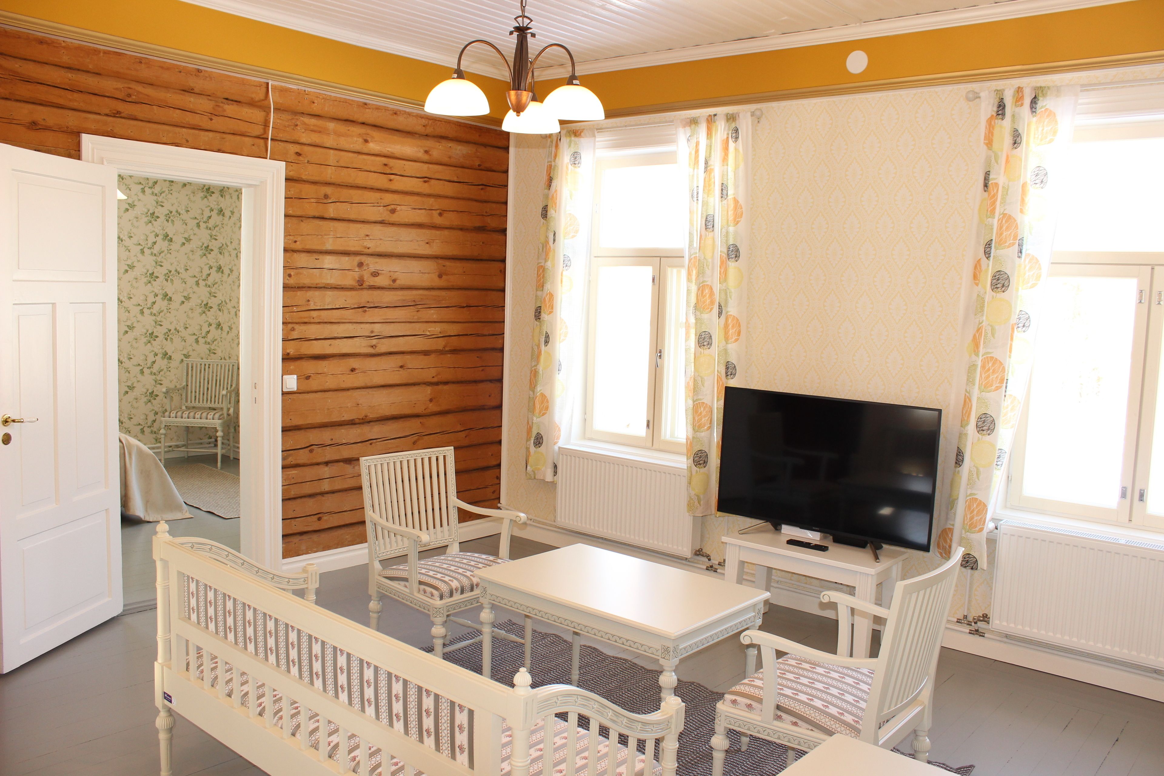 Una habitación con pared de madera, una mesa blanca, dos sillas blancas, un televisor y una ventana con cortinas.