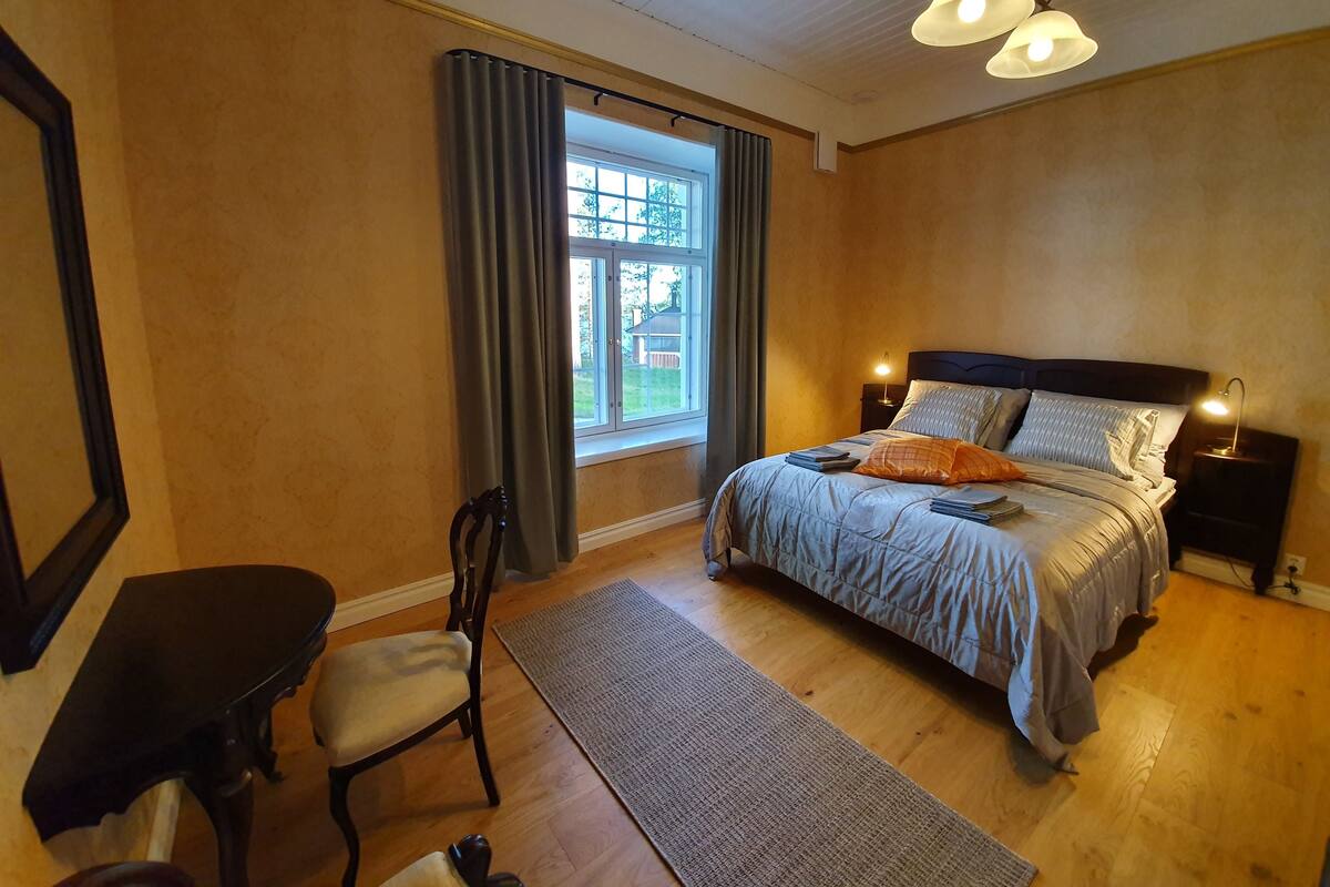 Een slaapkamer met een bed, een bureau, een stoel, een raam met gordijnen en een plafondlamp.