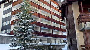 A multi-story building with balconies, a snow-covered tree, and a wooden building adjacent to it.