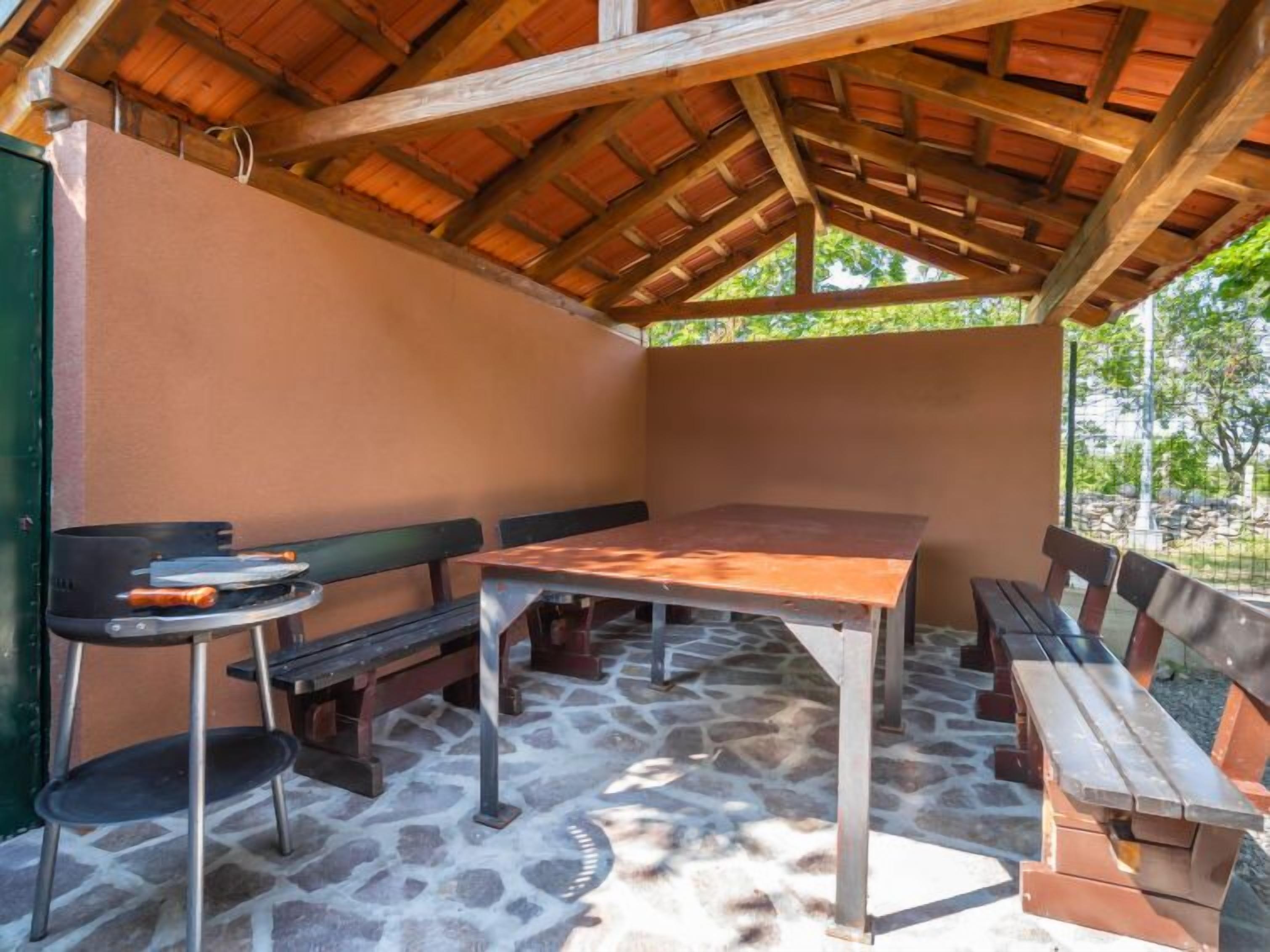 A covered outdoor seating area with a table and benches, stone flooring, and a wooden ceiling.