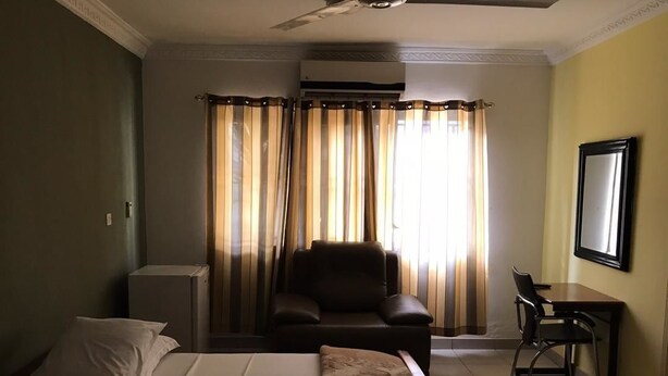 A hotel room with a bed, a desk, a chair, and a large window with curtains. - Mace Lodge (Sekondi-Takoradi)
