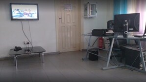 A room with a television mounted on the wall, a desk with a computer and a chair, and a cabinet with a sign above the door. - Mace Lodge (Sekondi-Takoradi)