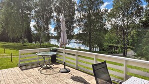 Une terrasse en bois avec une table et des chaises en verre, donnant sur un lac et entourée d’arbres.