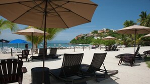 A beachside area with lounge chairs and umbrellas, a sandy beach, and a view of the ocean and buildings in the background. - Beautiful Condo in Alamar with fabulous OCEAN VIEW (La Cruz de Huanacaxtle)