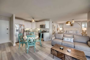 Une cuisine moderne avec des meubles blancs, un coin repas avec des chaises de couleur turquoise et un salon avec un canapé gris et une table basse.