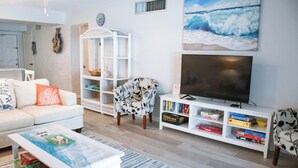 A living room with a white sofa, a patterned armchair, a coffee table, a bookshelf with books and games, a flat-screen TV, and a painting of a beach scene.