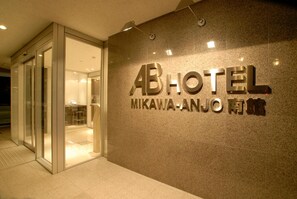 L’entrée d’un hôtel, avec une porte en verre et une enseigne indiquant « AB HOTEL MIKAWA-ANJO ».