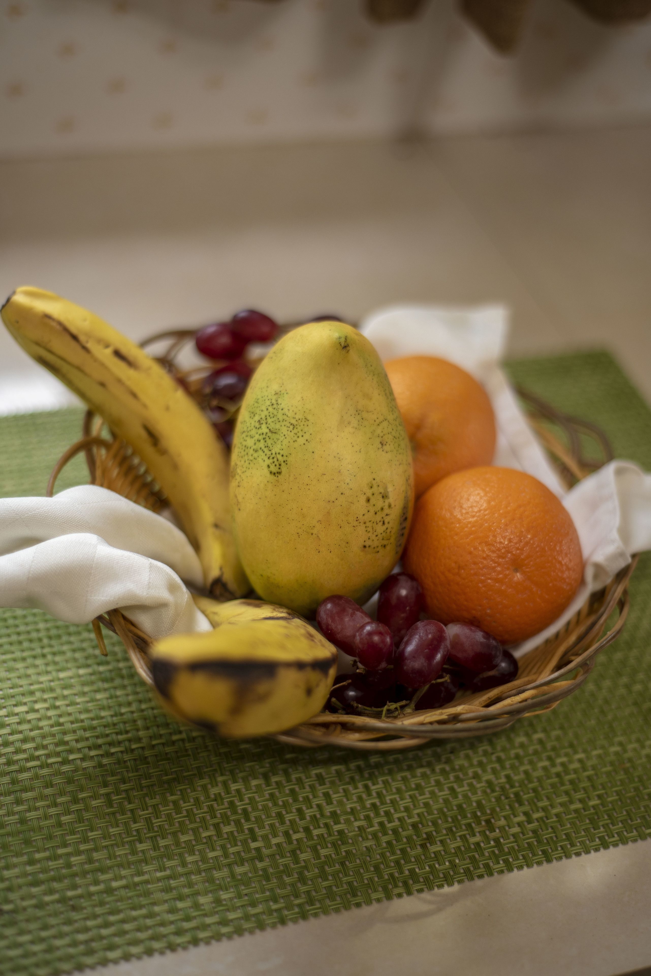 A wicker basket containing bananas, grapes, and an orange on a green mat.