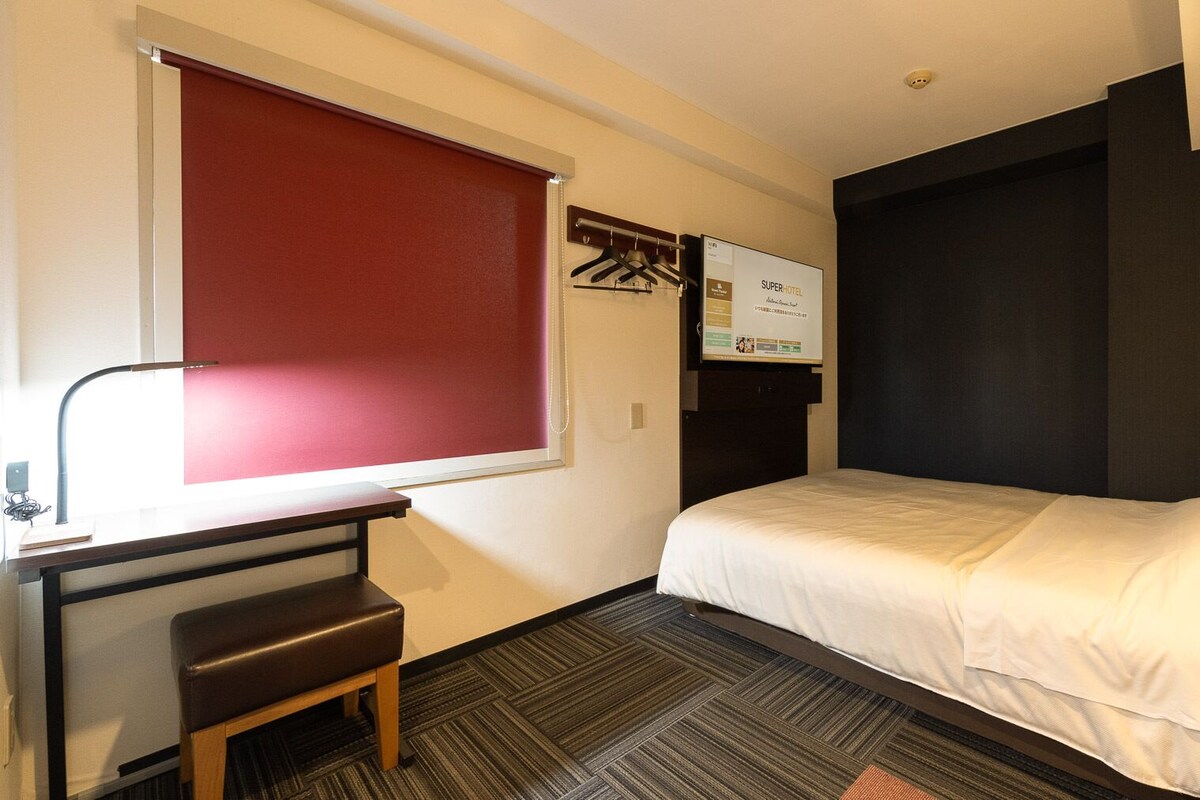 Een hotelkamer met een bed, een bureau met stoel, een raam met rood gordijn en een aan de muur bevestigde televisie.