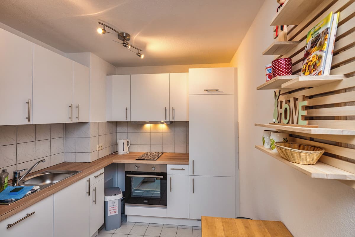 A compact kitchen with white cabinets, a built-in oven, a sink, and wooden shelves with various items.