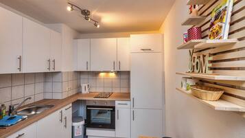 Una cucina compatta con mobili bianchi, forno integrato, lavandino e mensole in legno con diversi oggetti.