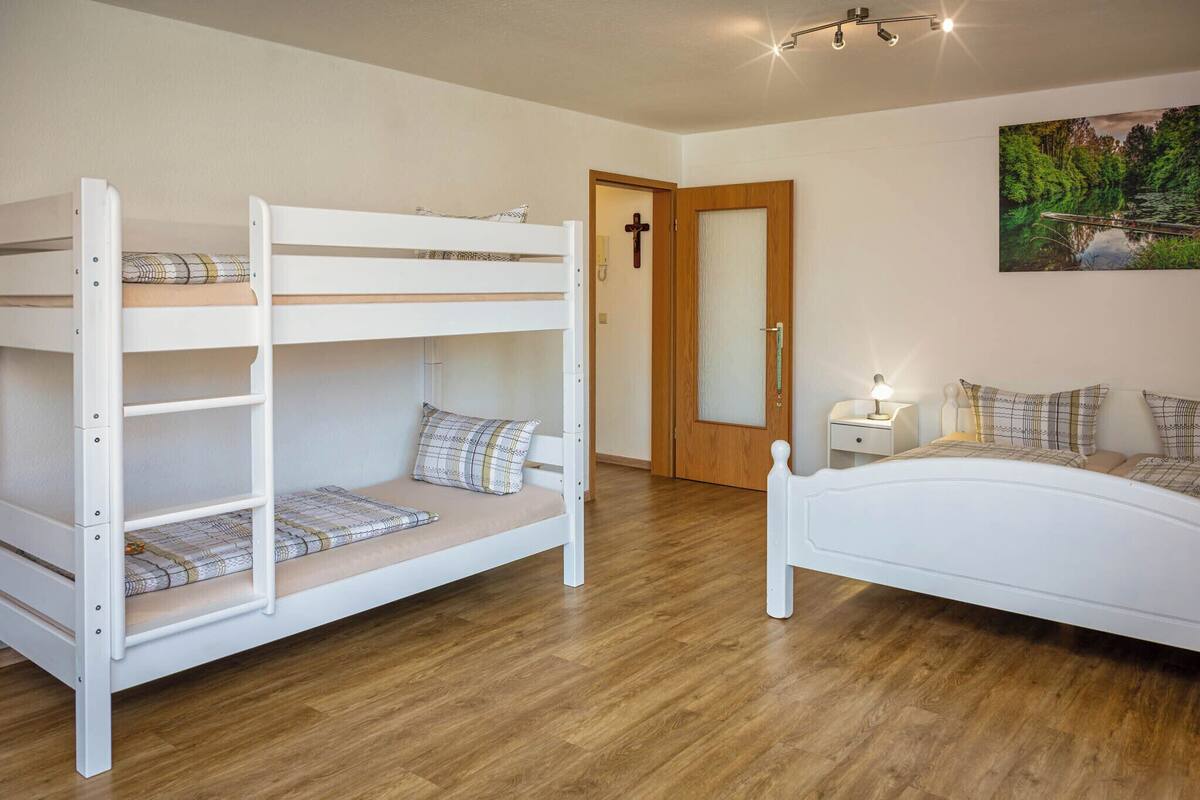 A bunk bed room with a wooden floor, a bedside table with a lamp, and a door with a cross.