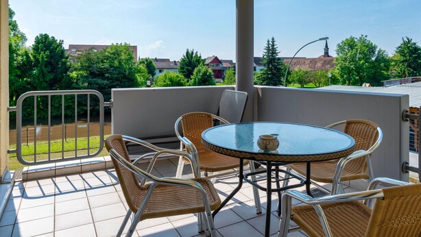 A covered outdoor area with a round table and four wicker chairs, a metal railing, and a view of a residential area with trees and houses. - Ferienwohnungen beim Europapark Rust (Rust)