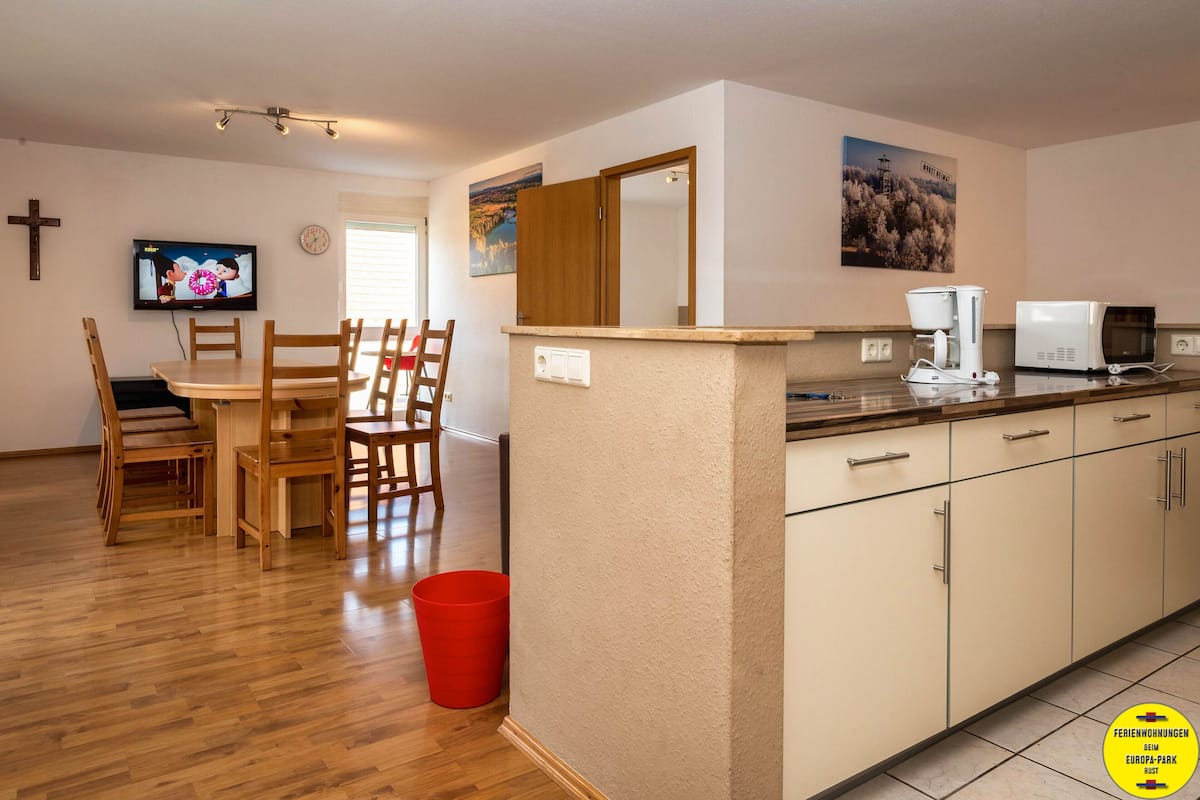 A modern kitchen with a wooden dining table and chairs, a red trash bin, and a television displaying a cartoon.
