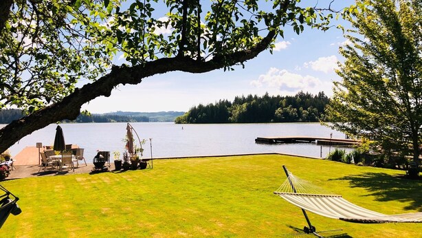 Um gramado com uma rede, um terraço com móveis de exterior e vista para um lago e uma floresta.
