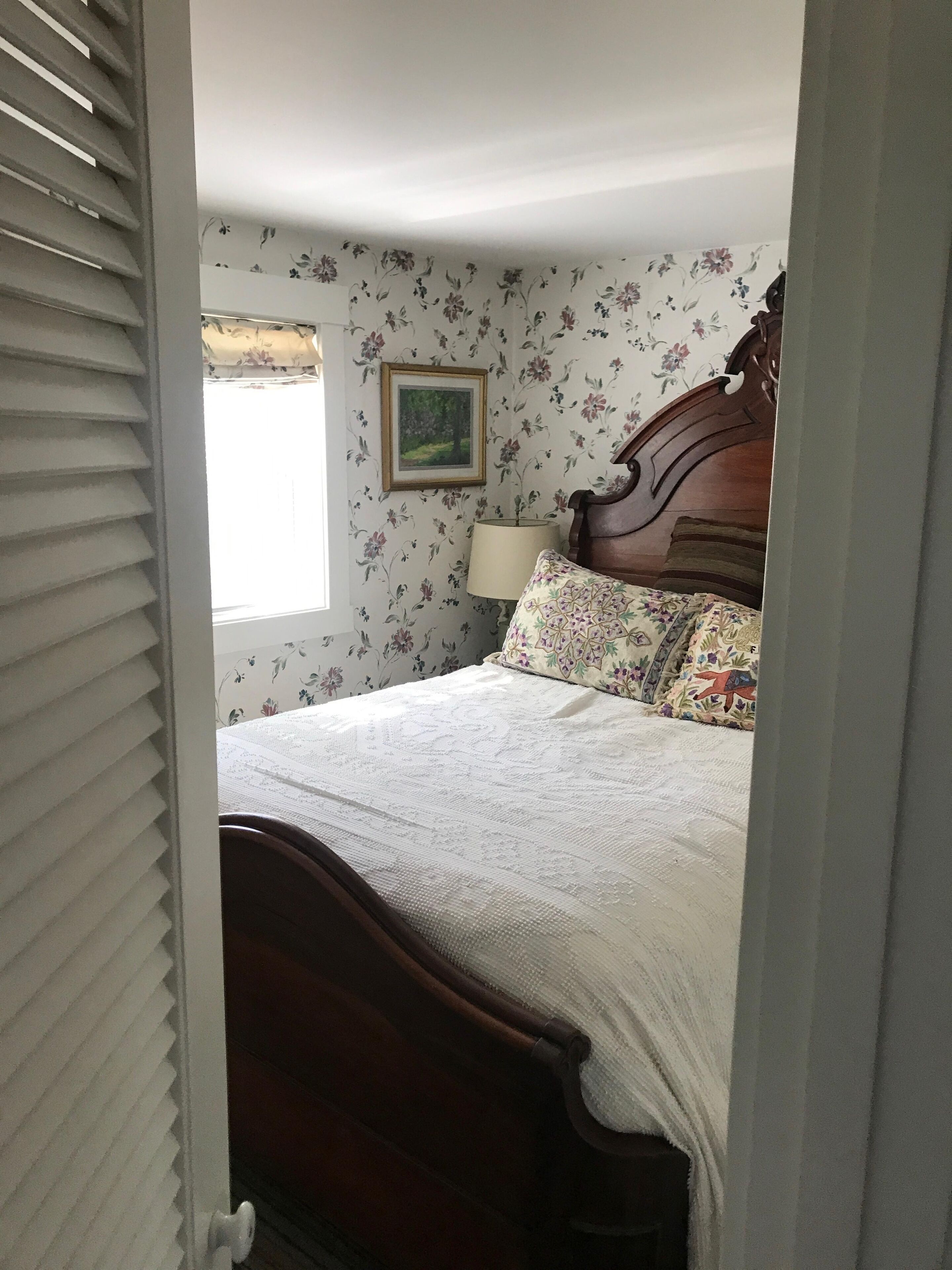 A bedroom with a wooden bed, floral wallpaper, a window with blinds, and a framed picture on the wall.