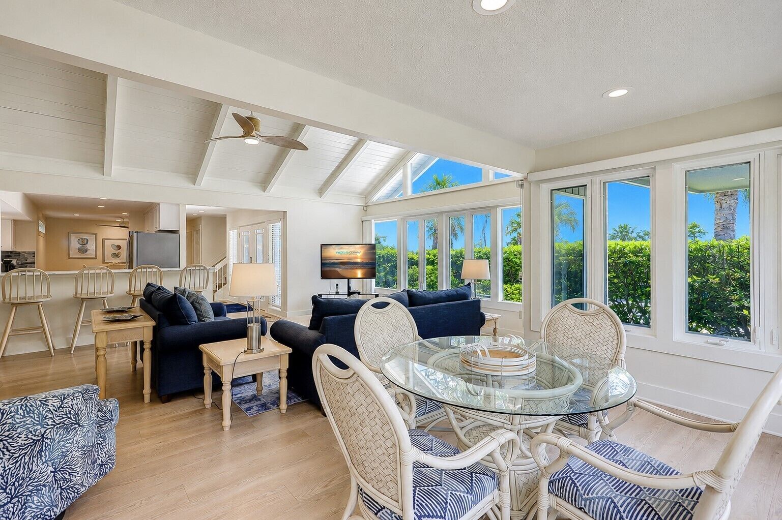 A spacious living area with a glass dining table surrounded by wicker chairs, a dark blue sofa, and a television on a stand.