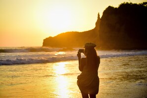 Eine Person macht bei Sonnenuntergang am Meer ein Foto, im Hintergrund sind Klippen zu sehen.