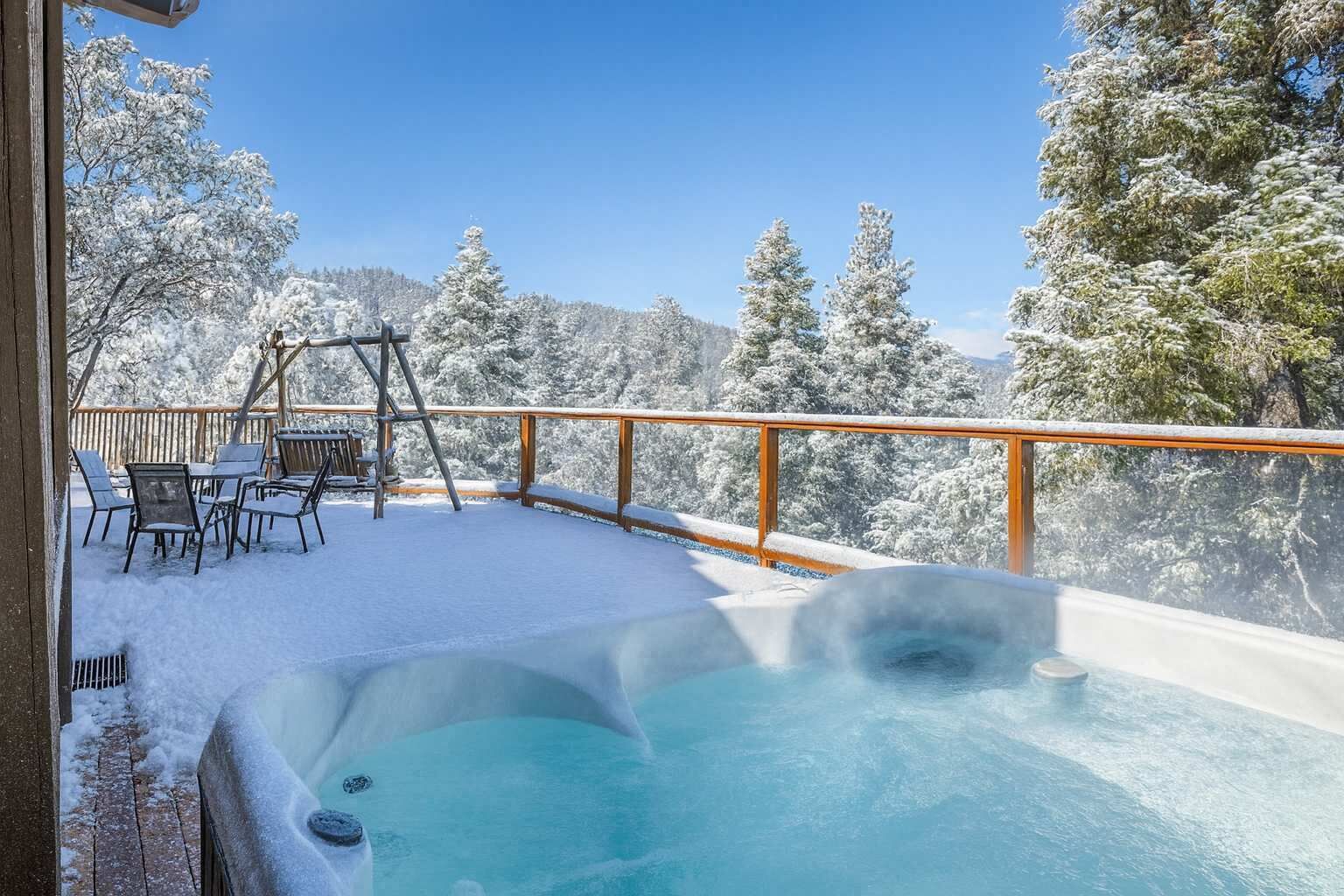 A snow-covered outdoor area with a hot tub, wooden deck, and chairs.