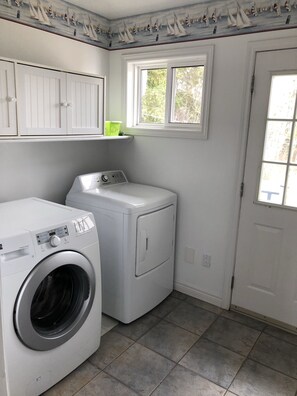A laundry room with a front-loading washer and dryer, a window with a view of greenery, and white cabinetry above the appliances. - Malagash Oceanside Vacation Home (Malagash Point)