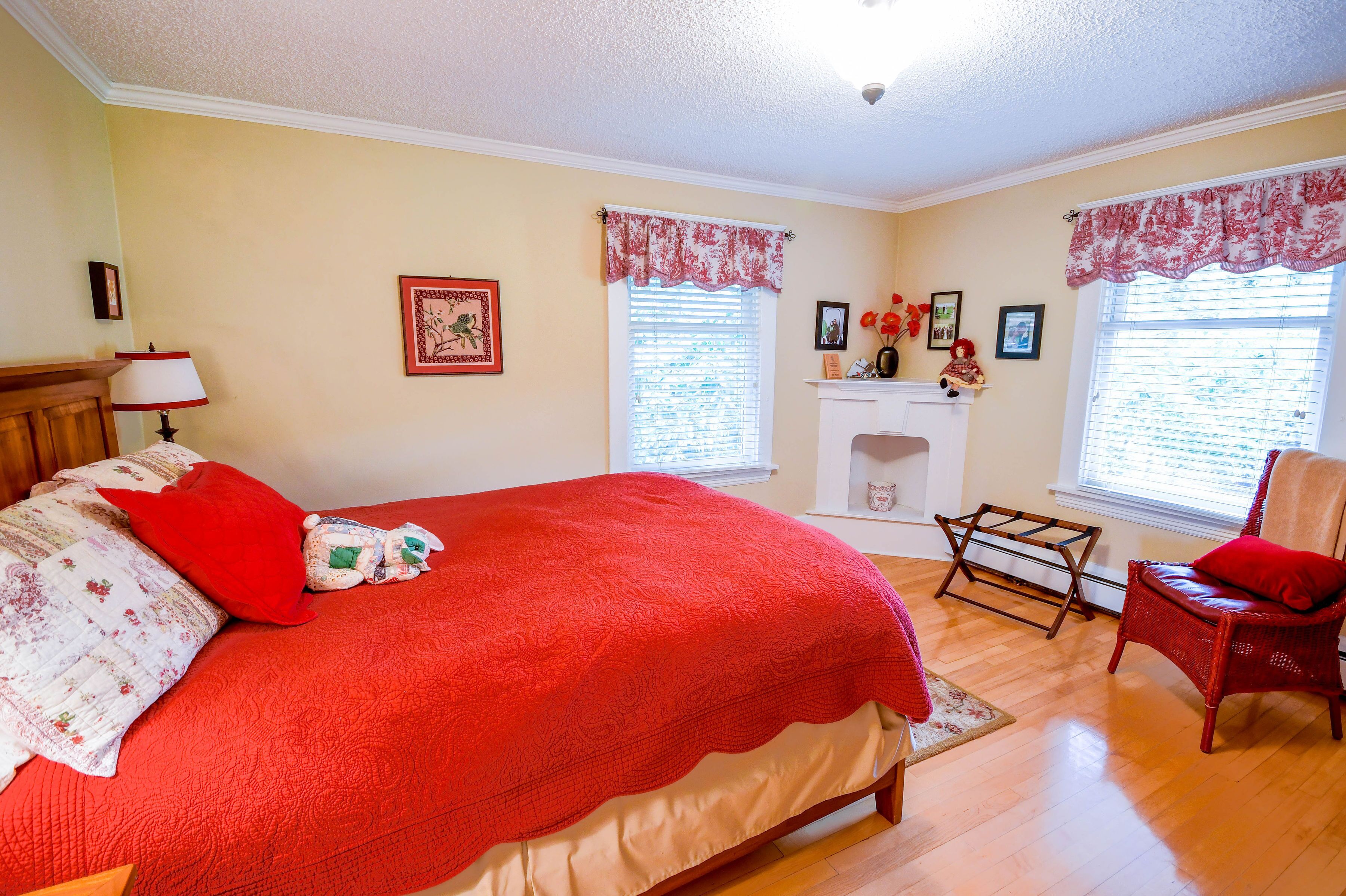 A bedroom with a large bed, two windows with shutters, a fireplace, and a chair.