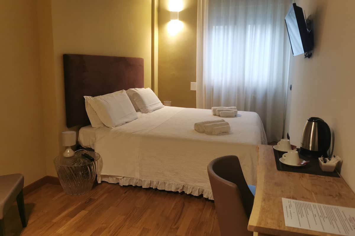 A hotel room with a bed, bedside table, chair, and a desk with a kettle and cup.