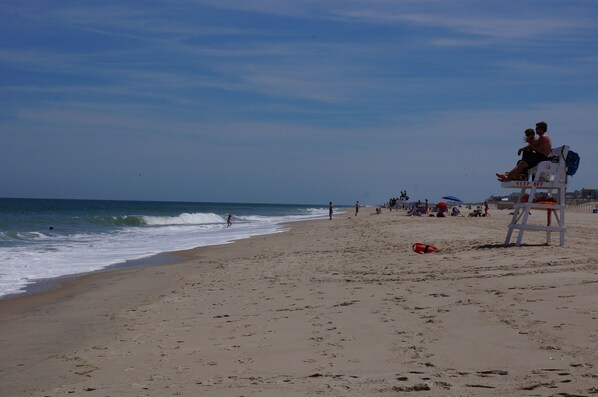 Una playa con una torre de salvavidas, gente caminando y sombrillas.