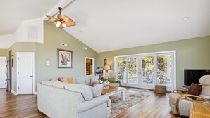 A spacious living room with a grey sectional sofa, a wooden coffee table, and a large sliding glass door leading to an outdoor area. - The Sand Trap in South Bethany: Steps from the ocean!  Hot Tub! Captain's walk! (Bethany Beach)