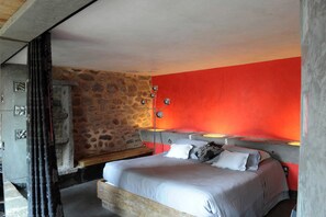 A bedroom with a stone wall, a red accent wall, a bed with pillows, a bedside table, and a lamp. - La Ferme de Moulhac (Laguiole)
