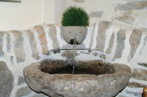 Une fontaine en pierre, surmontée d’un pot contenant une plante, le tout adossé à un mur de pierre.
