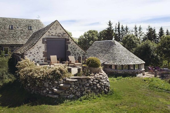 A stone house with a sloped roof, a covered patio with outdoor furniture, and a stone wall with a small bush. - La Ferme de Moulhac (Laguiole)