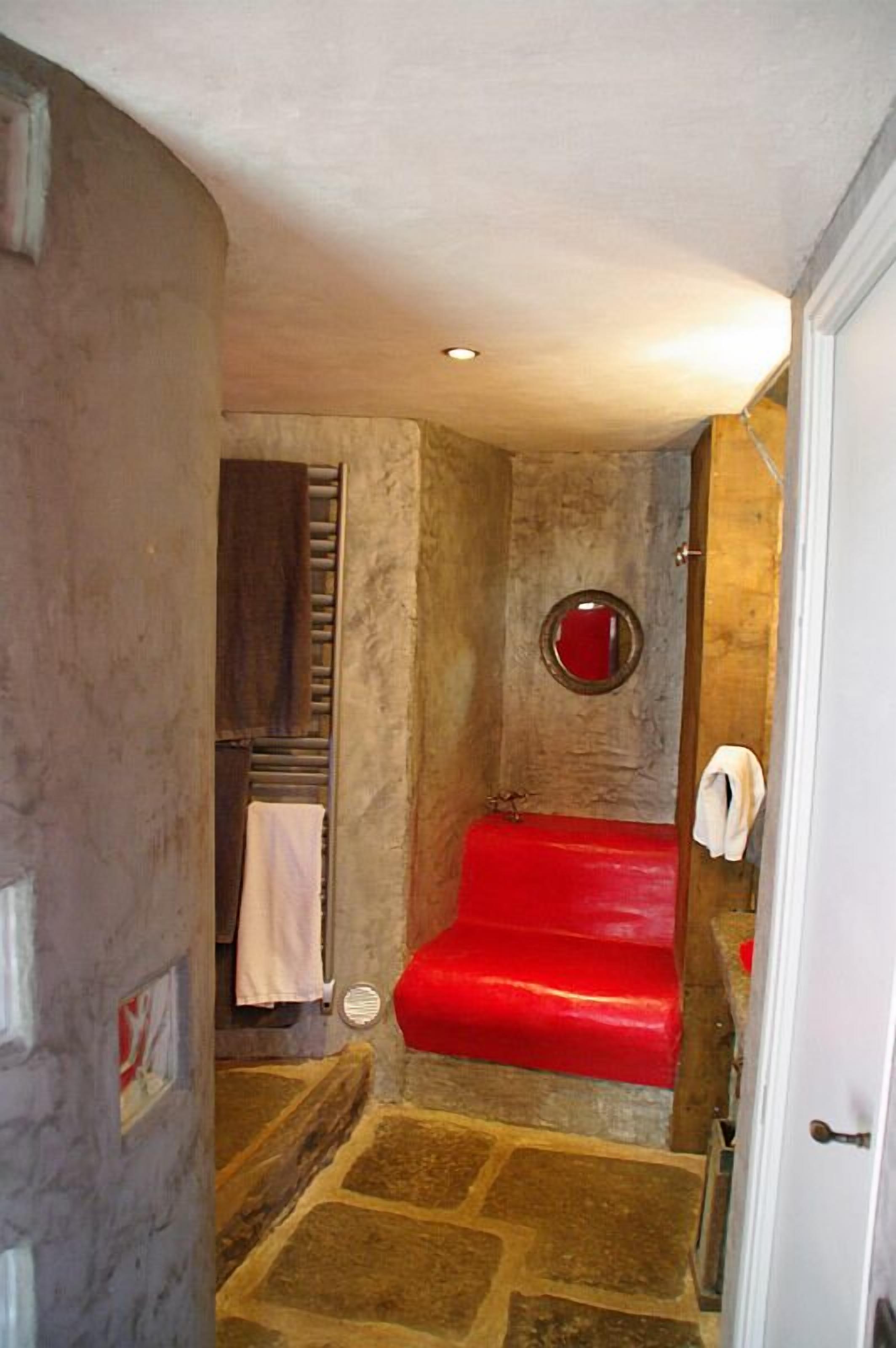 A bathroom with a stone floor, a red bathtub, a small round mirror, and a towel hanging on the wall.