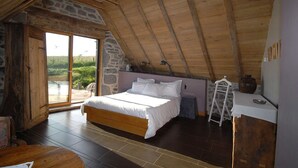 Une chambre avec un plafond en bois, des murs en pierre, un grand lit, une petite table et une vue sur l’extérieur, visible à travers une porte vitrée.