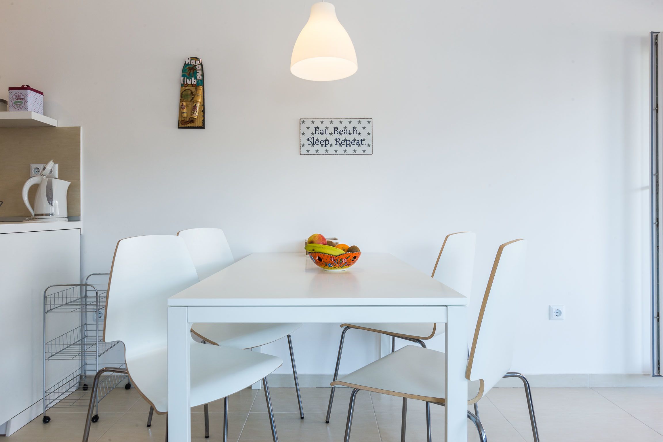 Un espace repas moderne avec une table blanche, quatre chaises, un saladier de fruits, une lampe suspendue et un panneau fixé au mur.