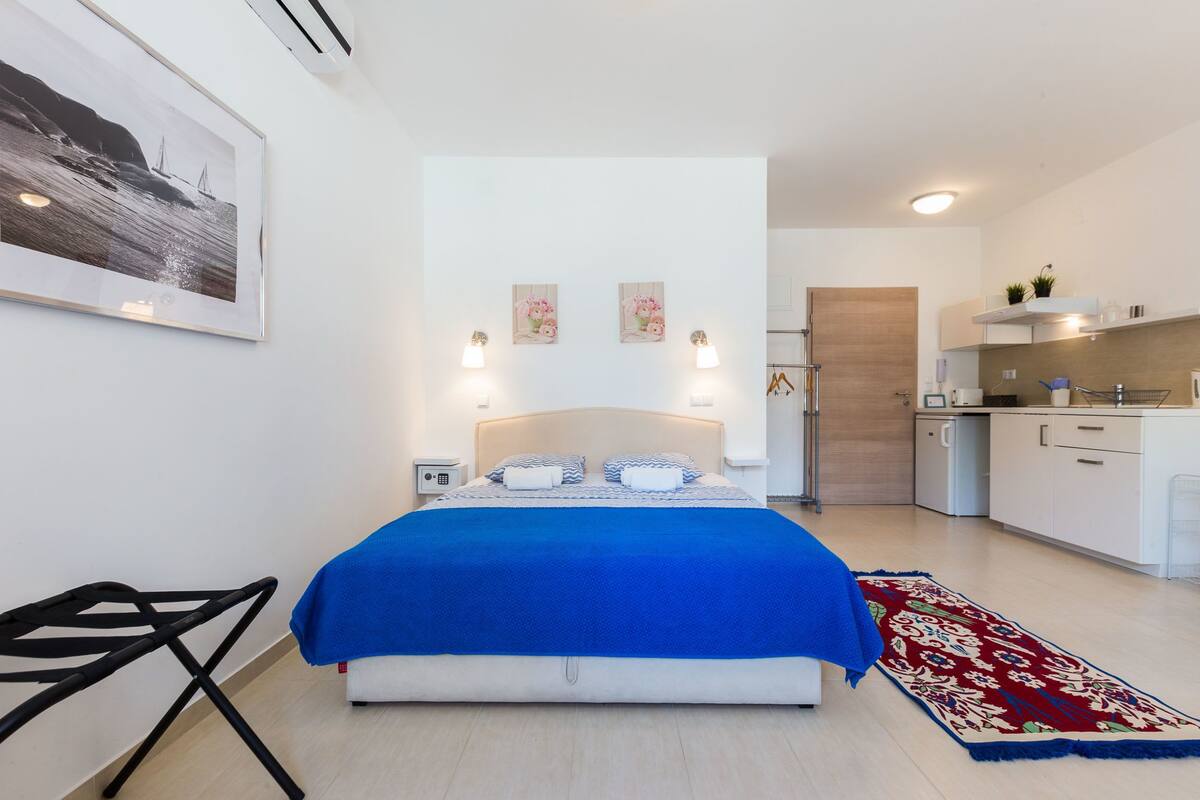 A modern bedroom with a large bed, bedside tables, a small desk, and a view into the kitchen.