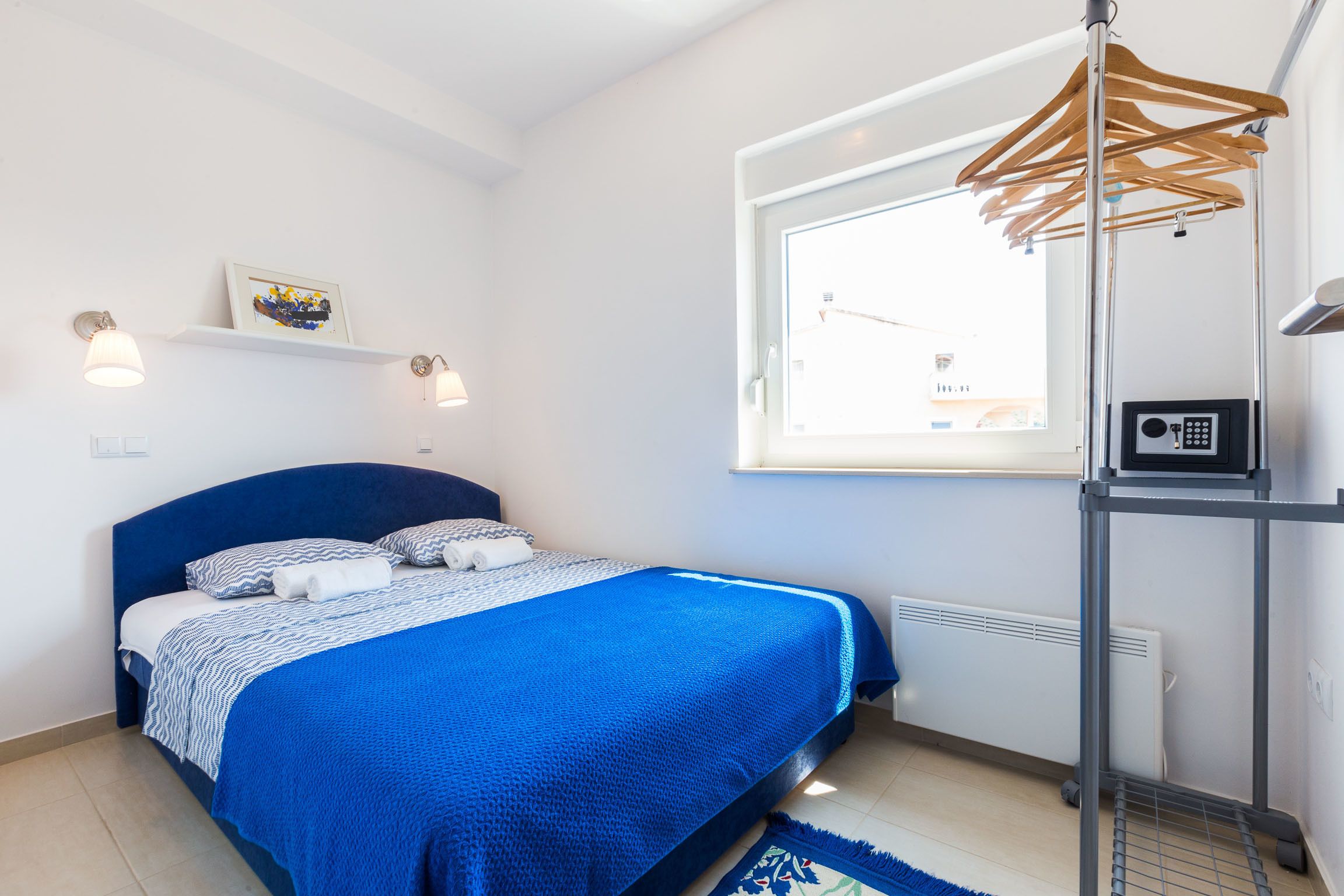 A bedroom with a bed, a window, a radiator, and a shelf with a picture.