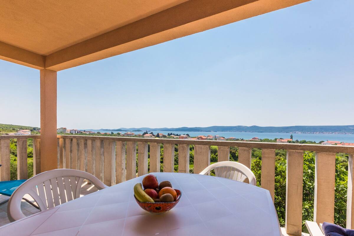 A table with a fruit bowl on a balcony overlooking a scenic view.