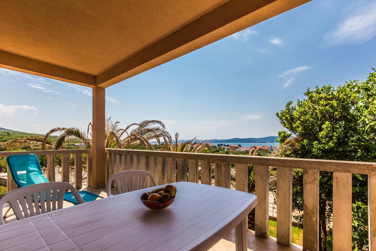 A covered outdoor dining area with a table and chairs, a bowl of fruit, and a view of the landscape.