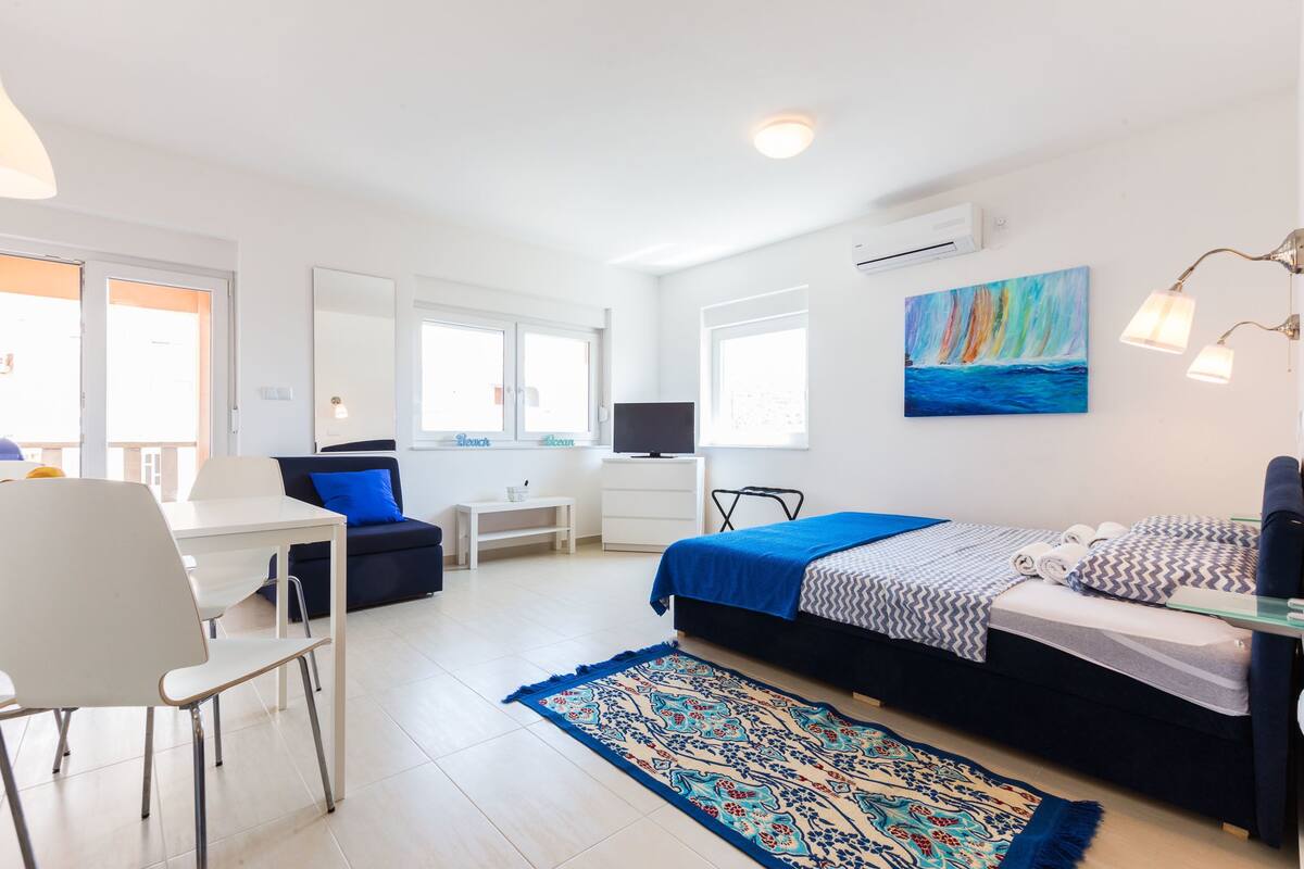 A modern bedroom with a bed, a desk, a chair, a TV, and a painting on the wall.