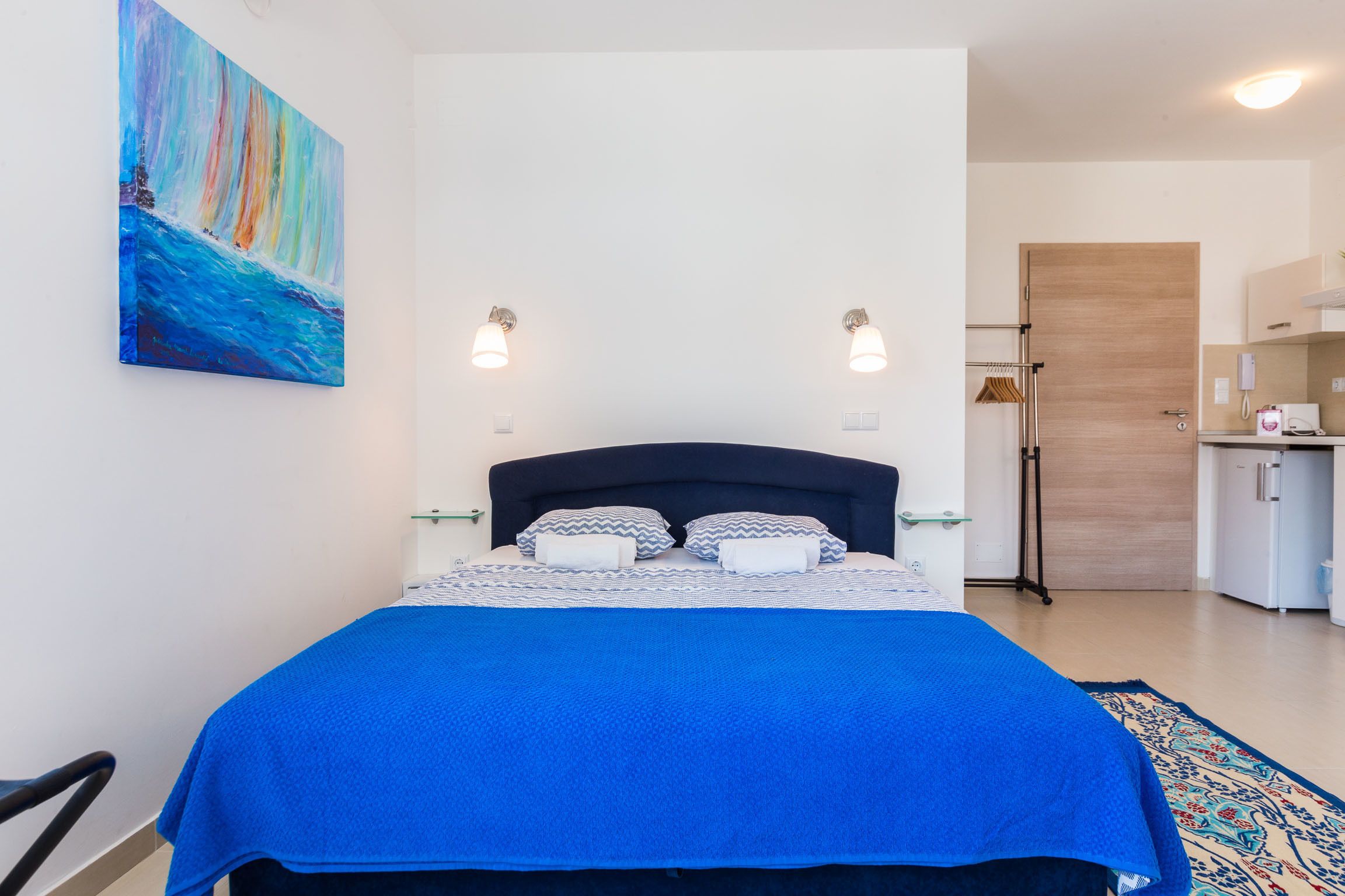 Un dormitorio con cama, mesita de noche, un cuadro y una kitchenette.
