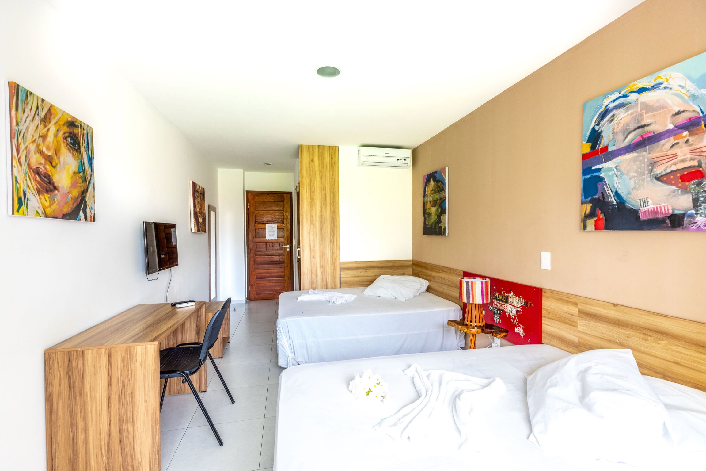 Ein Hotelzimmer mit zwei Betten, einem Schreibtisch und einem Stuhl. An den Wänden befinden sich Gemälde und eine Klimaanlage.