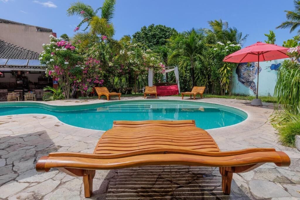 A swimming pool with wooden lounge chairs and a red umbrella, surrounded by lush greenery and a mural.